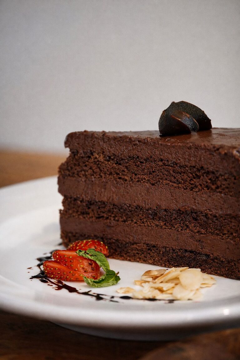 Close view of a chocolate layer cake slice with glossy ganache, flaked almonds and a fanned strawberry.