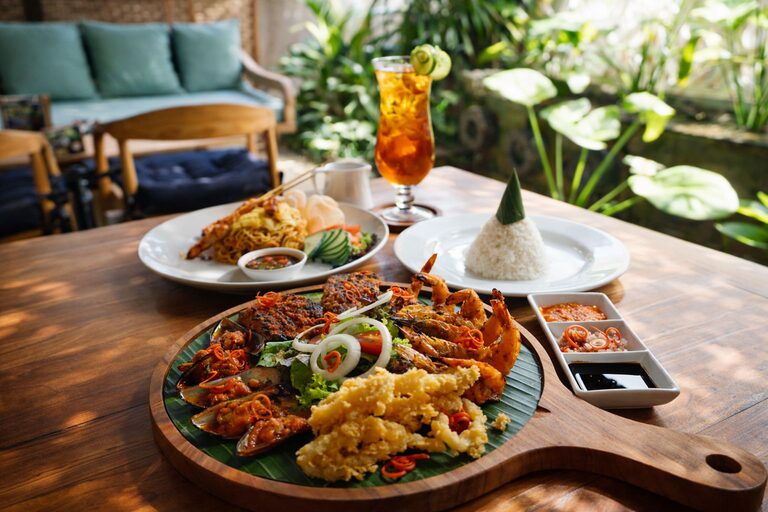 A seafood platter on a banana leaf with grilled prawns, mussels in sambal, fish and calamari, set on a sunlit teak table.